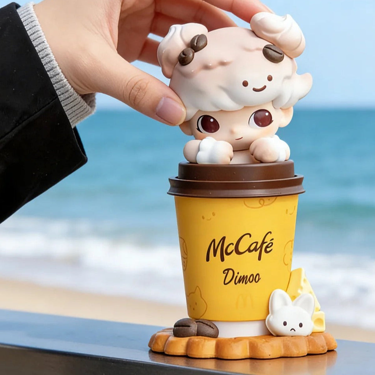 DIMOO McCAFE — cup-shaped figure shown by hand in a beach display scene