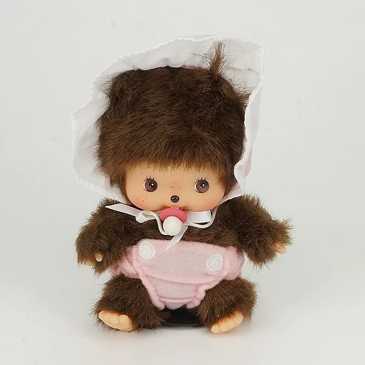 Monchhichi D plush doll wearing a pink bib and white bonnet, seated front view