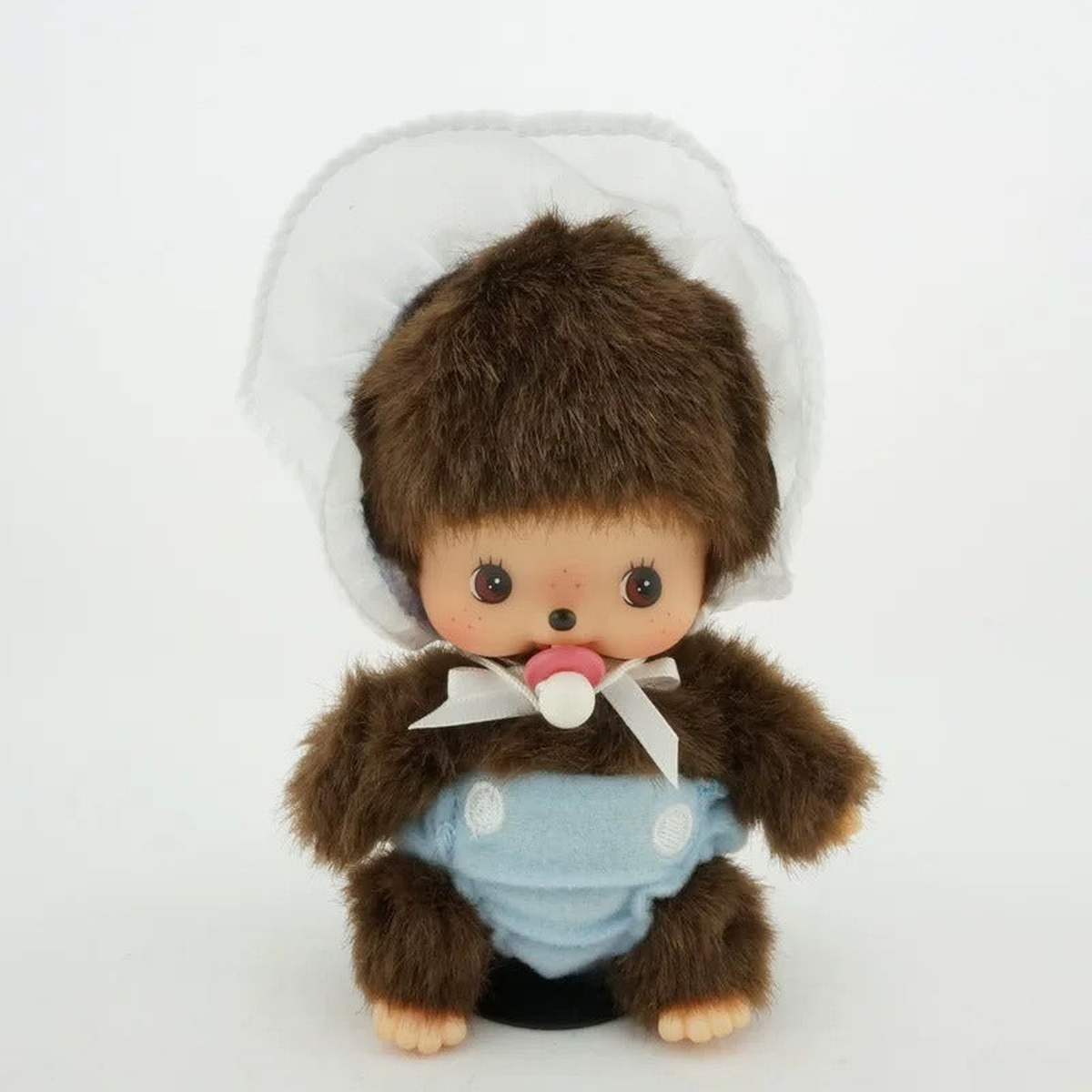 Monchhichi E plush doll wearing a light blue bib and white bonnet, seated front view