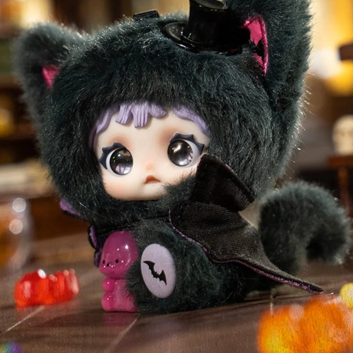 NOMMI x HEYMI dark plush doll with a small top hat, bat belly patch and pink bear ornament in an indoor scene
